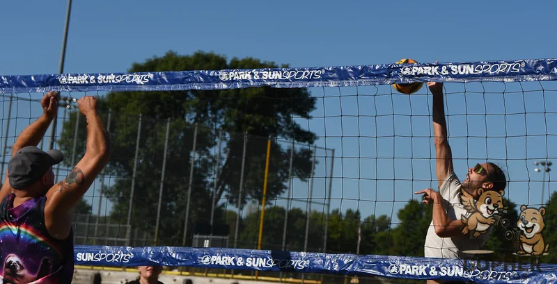 A beach volleyball player reaches for the ball above the net while an opponent raises both hands to block.